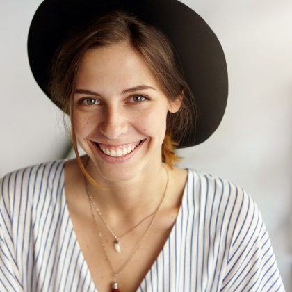 Waist up portrait of beautiful European female with dark eyes, warm smile and dark hair wearing black hat and blouse posing in camera having good mood. Smiling elegant young attractive woman in hat