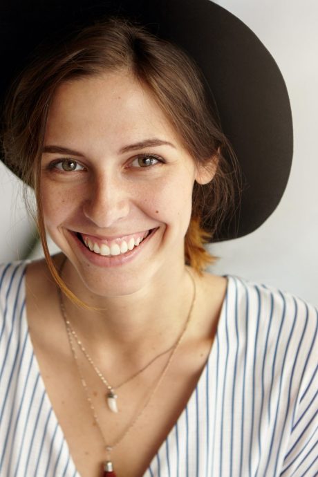 Waist up portrait of beautiful European female with dark eyes, warm smile and dark hair wearing black hat and blouse posing in camera having good mood. Smiling elegant young attractive woman in hat